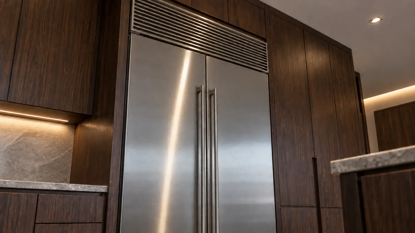 A built-in refrigerator standing flush with the surrounding cabinetry, its ventilation grille running across the top above the doors.